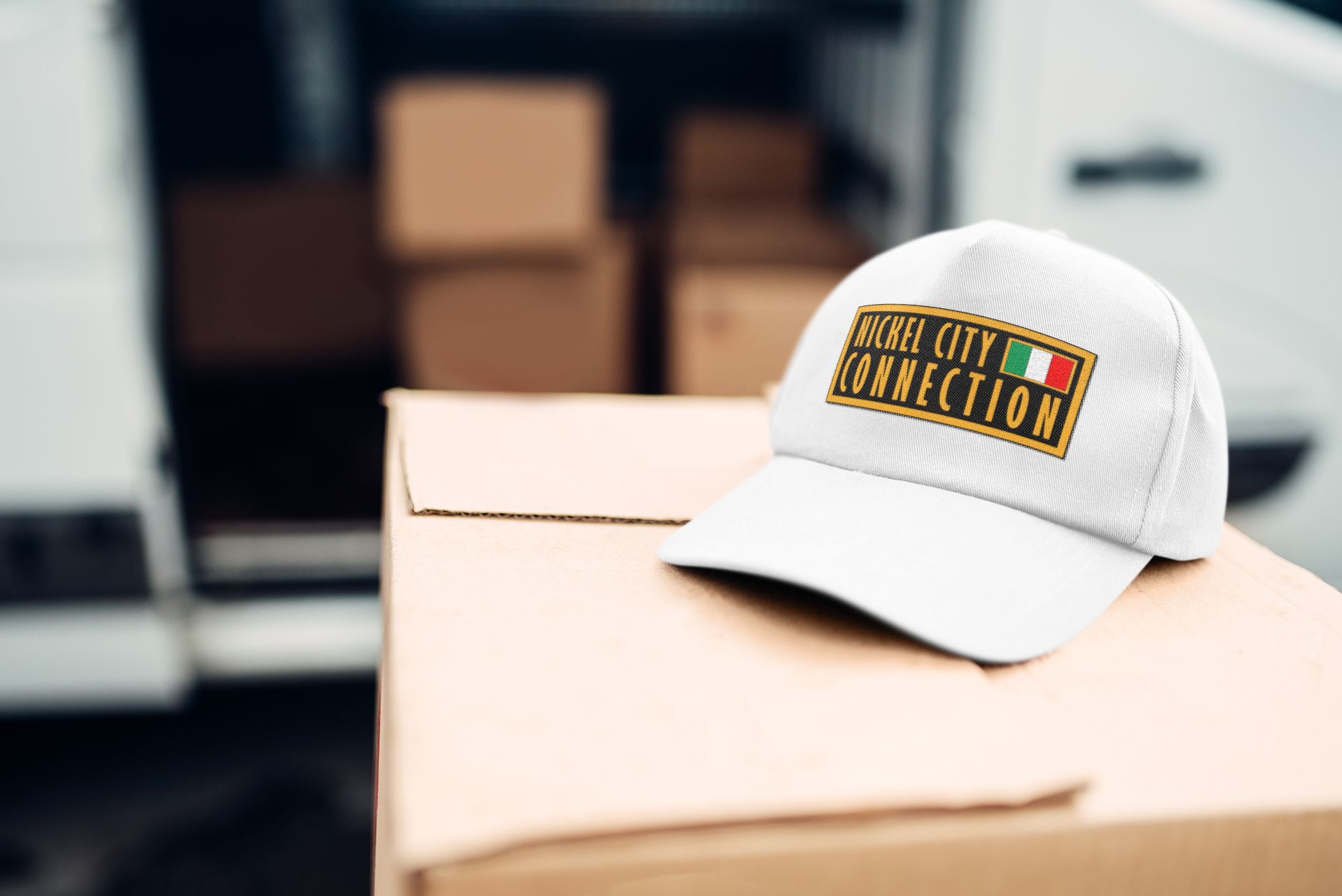 A white "Nickel City Connection" hat rests on a cardboard box, with a blurred background of stacked packages. "Hats" and a "Shop" button overlay the image.