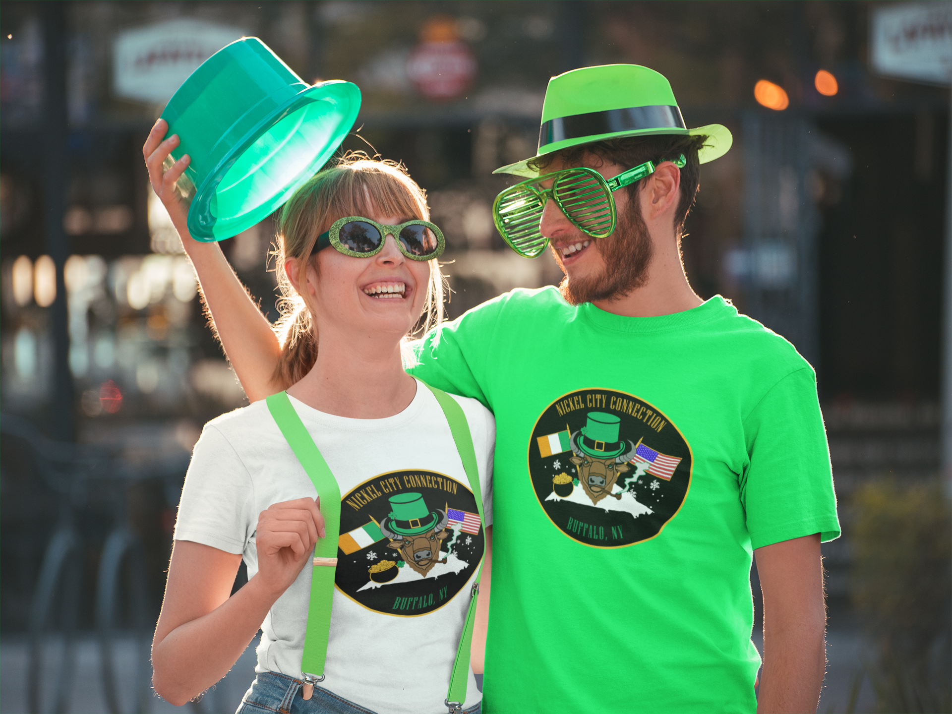 A cheerful couple in festive Irish-themed outfits wears matching "Nickel City Connection" shirts featuring a bison in a leprechaun hat, Irish and American flags, and a pot of gold. The woman sports green suspenders and round glasses, while the man wears a green hat and shutter shades. "Irish American Pride Collection" text and a "Shop" button overlay the image.